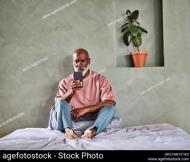 Mature bald man surfing net through mobile phone sitting on bed at home