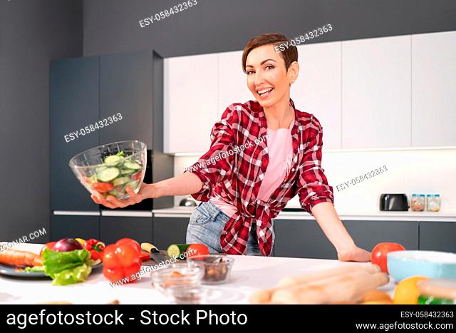 Happy holding bawl of salad young housewife cut tomato, cucumber, pepper for a family dinner or girls night standing in the kitchen