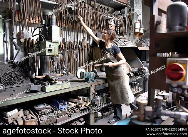 27 July 2023, Saxony, Hohenprießnitz: 39-year-old blacksmith and metal designer Marika Widdermann fetches forging tongs from a rack in her father Roger...