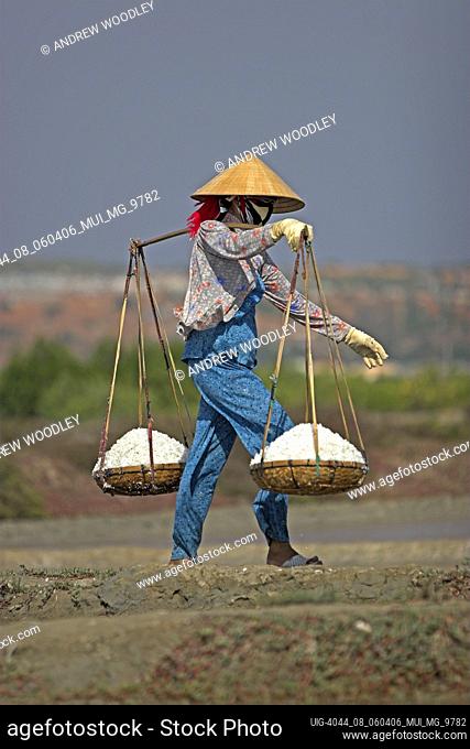 Woman in conical hat carrries wicker pannier baskets of salt between salt ponds Phan Thiet Vietnam