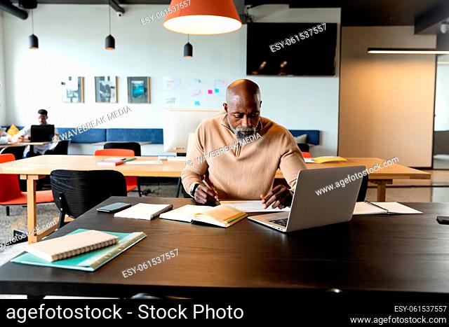 African american mature businessman writing in diary while sitting with at laptop in creative office