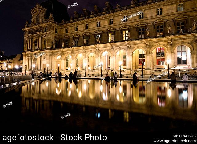 Tourists sit in front of the Brasserie Le Café Marly in the Musée du Louvre at night and reflect themselves in the water, Paris, France, Europe