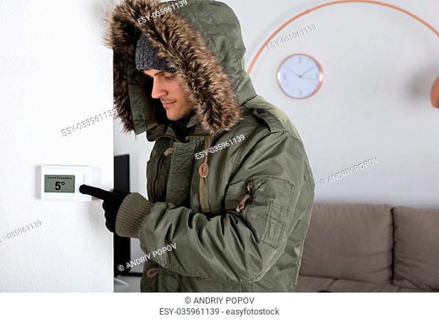 Young Man In Warm Clothing Pointing To Current Room Temperature In Digital Thermostat At Home