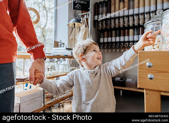 Cheerful boy holding hand of father while pointing at candy jar in store