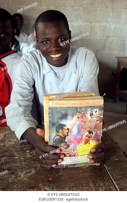 Tanji village. Smiling student from Usman Bun Afan private Islamic school proudly showing his exercise book with picture of a football referee showing a yellow...