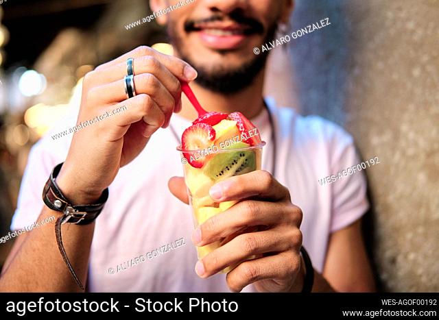 Smiling man holding fruit salad in disposable glass while standing by wall