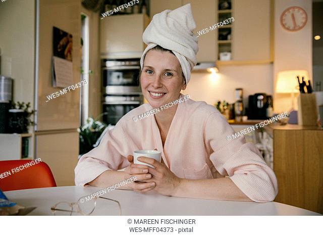 Portrait of smiling woman wearing a towel turban sitting with cup of coffee at table in the kitchen