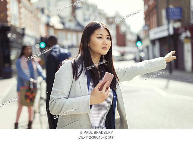 Young businesswoman with cell phone hailing taxi on sunny urban street
