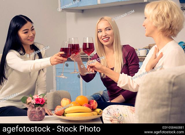 Toned picture of happy friends girls drinking red wine and smiling while spending free time at home. Beautiful cheerful ladies having party in kitchen after...