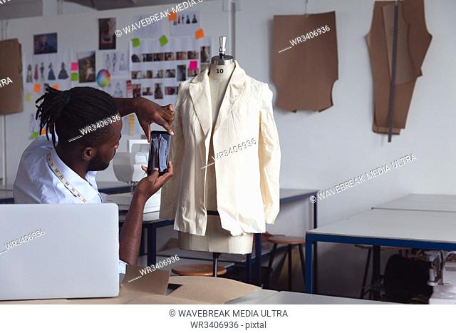 Side view of a young African American male fashion student using a smartphone to take a photo of a jacket design on a mannequin in a studio at fashion college