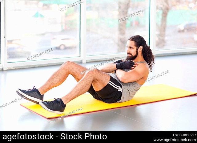 Portrait of young adult muscular built athlete with long curly hair working out in gym, lying on mat and swinging press with crossed arms on chest and bent legs