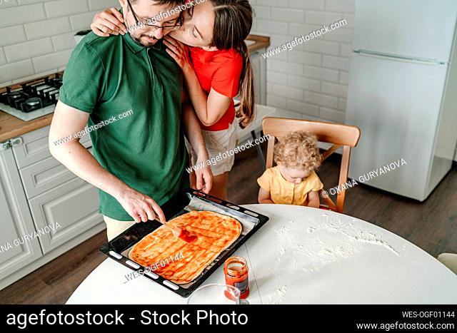 Woman kissing man preparing pizza by daughter sitting on chair in kitchen