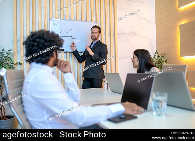 Corporate manager pointing with a marker at the horizontal line on the graph on the whiteboard to his colleagues