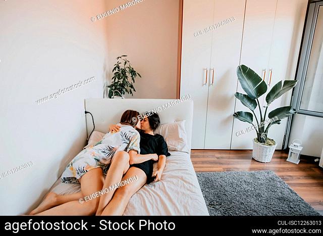 Two women with brown hair lying on white daybed, hugging and kissing