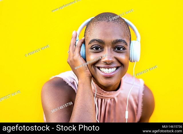 close up and portrait of a young afro american woman looking at the camera smiling and enjoying music with her headphones - yellow background