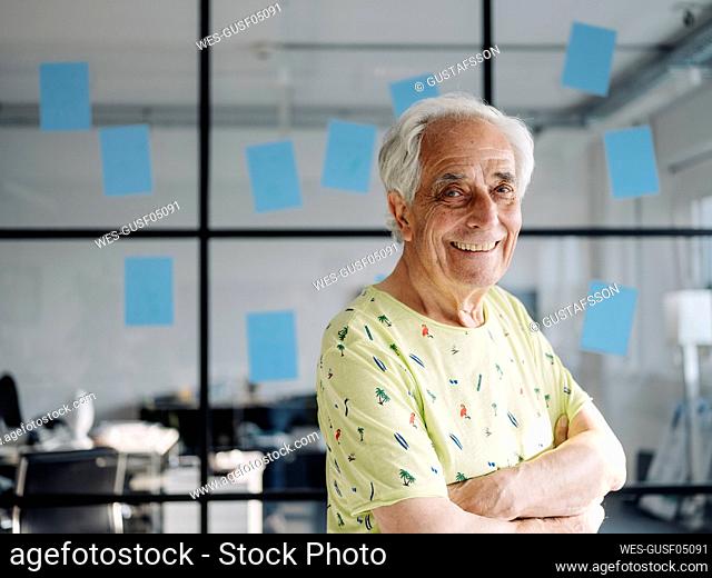 Confident businessman with arms crossed smiling while standing against glass wall