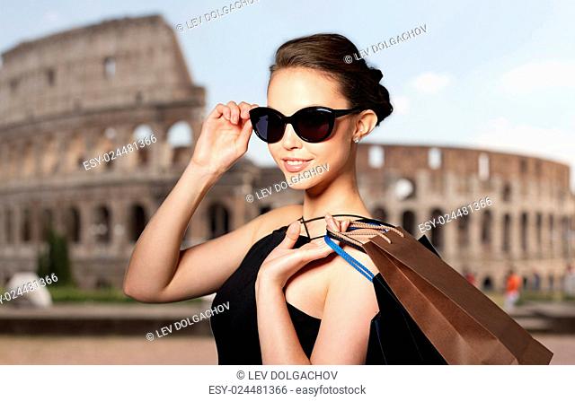 sale, tour, fashion, people and luxury concept - happy beautiful young woman in black sunglasses with shopping bags over coliseum background