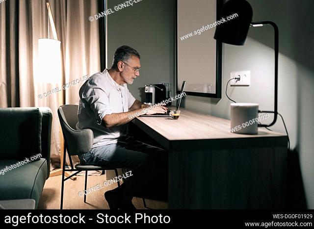 Mature man sitting at desk with coffee while using laptop in hotel room