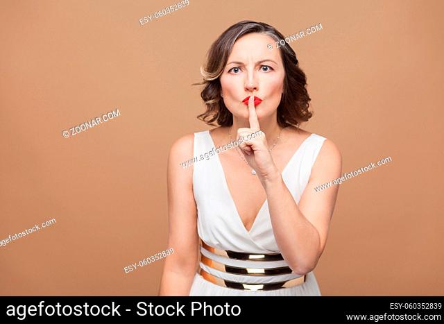 bossy middle aged woman showing shh secret sign. Emotional expressing woman in white dress, red lips and dark curly hairstyle