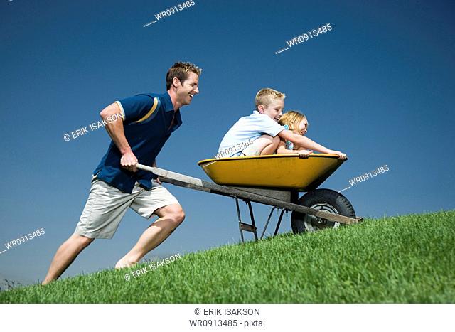 Father pushing son and daughter in a wheelbarrow