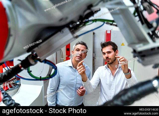 Young male engineer discussing over robotic machinery with colleague in industry