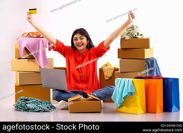 A young woman sitting with laptop and credit card amidst cardboard boxes, clothes and colorful shoppingbags