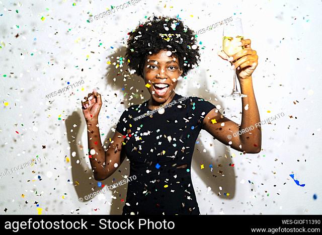 Happy woman with champagne flute dancing while standing amidst confetti against white background