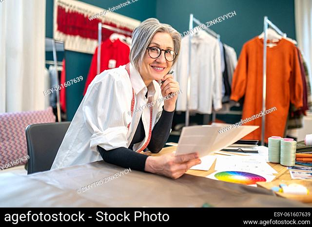 Working mood. Smiling middle aged woman in glasses sitting at table with work mess holding sketch looking at camera in atelier
