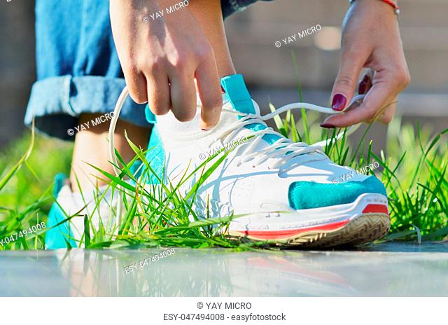 Young girl wearing jeans tying shoelaces on sneakers standing on green grass side view close-up horizontal photo, Sunny day