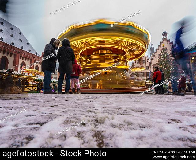 04 December 2023, Hesse, Frankfurt/Main: A classic carousel appears blurred by a long exposure. Snowfall transforms the Christmas market on the Römerberg into a...