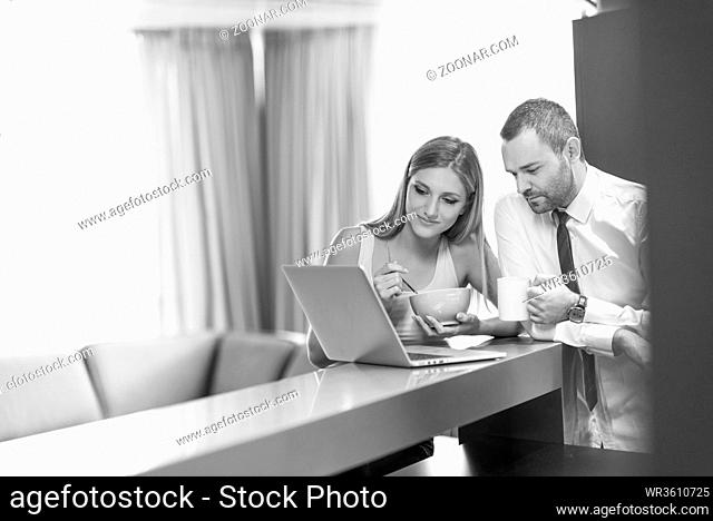 A young couple is preparing for the job and using a laptop. The man drinks coffee while the woman eats breakfast at luxury home together, looking at screen