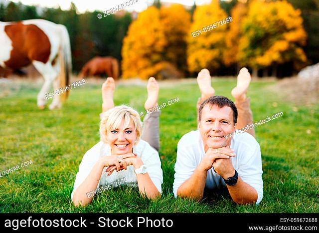 Couple lies on the lawn with their arms folded under their chins in the aurumn forest. Horses graze in the background. High quality photo