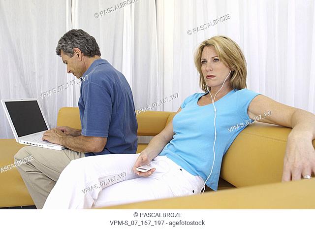 Mid adult woman listening to an MP3 player with a mid adult man using a laptop