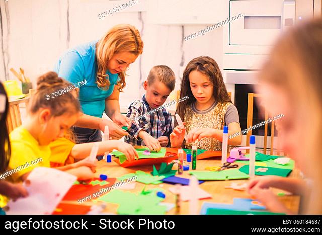 The preschoolers sit in a room at a table and make applications from colored paper. They are helped by an adult woman