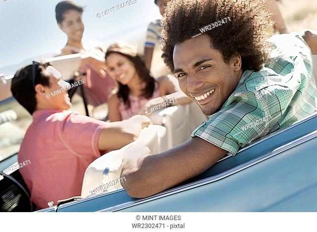 A group of friends in a pale blue convertible on the open road, driving across a dry flat plain surrounded by mountains