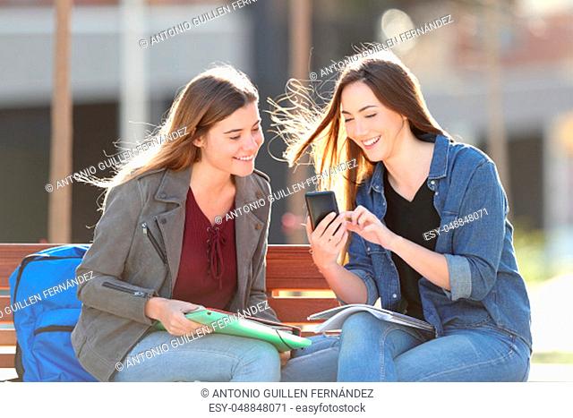 Two happy students checking smart phone content sitting on a bench in a park