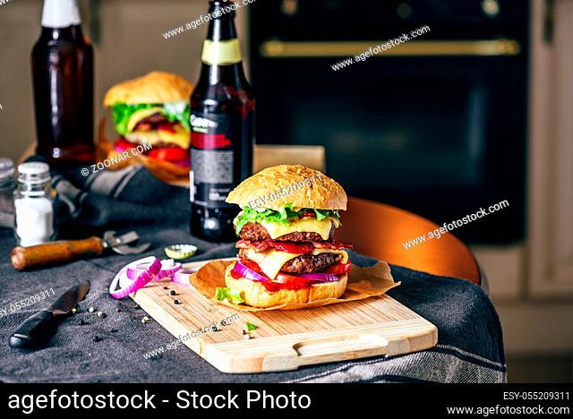 Burger with Two Beef Patties, Cheddar Cheese, Bacon, Iceberg Lettuce, Sliced Tomatoes and Red Onion on Cutting Board. Bottle of Beer and Some Ingredients on...
