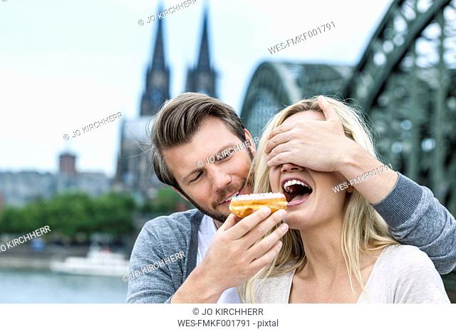 Germany, Cologne, young woman tasting a bagel while her boyfriend covering her eyes