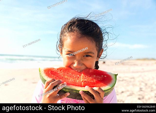 Portrait of cute biracial girl eating large fresh watermelon slice at beach on sunny day