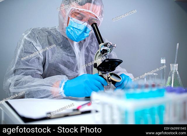 Medical professional in white protective suit looking through microscope in chemical laboratory. Search vaccine coronavirus. Covid 19 drug inventions