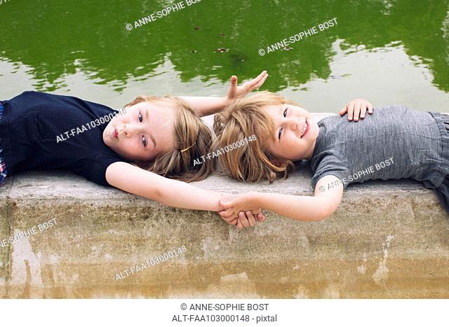 Little girls lying together beside pond, holding hands