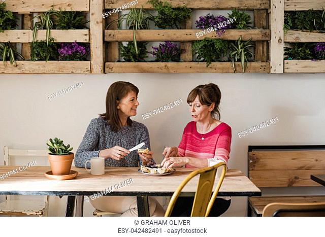 Two Female friends sitting inside a little cafe relaxing. They are buttering a scone and enjoying a coffee