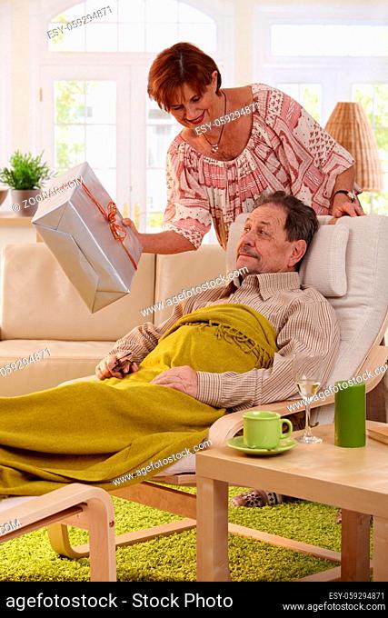 Senior couple celebrating birthday. Wife giving present to her husband, resting in armchair