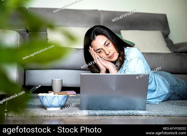 Serious woman looking at laptop while lying on carpet at home