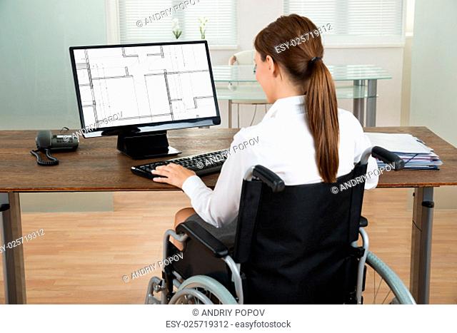 Young Female Architect On Wheelchair Looking At Blueprint On Computer In Office