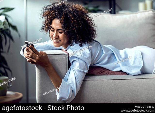 Happy woman text messaging over smart phone lying on sofa