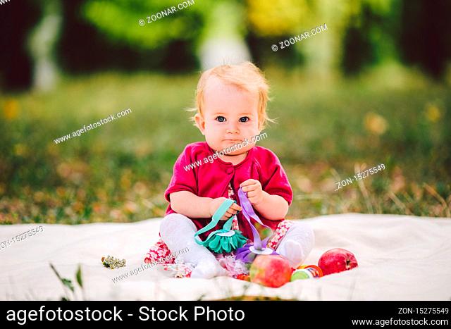 Little baby girl Caucasian ethnicity blond one year old from birth sits on a plaid on green grass in the park. Child at picnic in the autumn forest