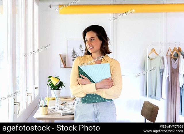 Smiling young female design professional holding file while looking away at clothing store