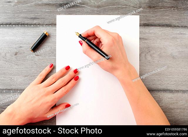 View from above to woman's hands with red nails, holding black fountain ink pen with gold nib, ready to write something on empty piece of paper laying on gray...