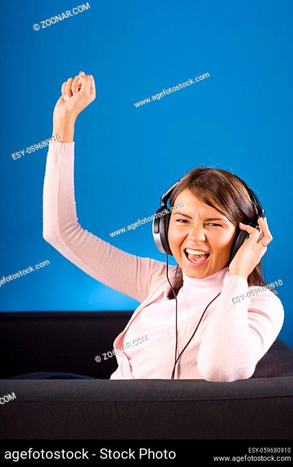 Young woman sitting on the black sofa. Wearing headphones; blue background, in studio